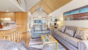 A modern living room with a sofa, coffee table, and kitchen area with wooden cabinets and a bar. - Golf Course Views, Half Mile to the Village - Includes SHARC Access (Sunriver)