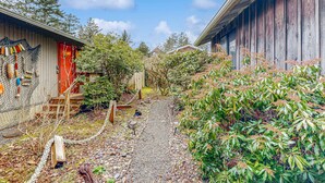A narrow pathway leading to a wooden building with a red door, surrounded by greenery and a fence. - Dog-Friendly Cottage with Private Hot Tub, Deck, Berry Bushes, & Beach Access (Cannon Beach)
