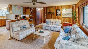 A living room with a sofa, a coffee table, a bookshelf, a map on the wall, and a window with a view. - Dog-Friendly Cottage with Private Hot Tub, Deck, Berry Bushes, & Beach Access (Cannon Beach)