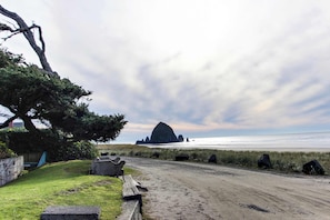 A coastal road with a grassy area, a wooden bench, and a large rock formation in the distance.