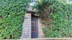 A small, dark-colored door with a window, surrounded by dense green foliage. - Dog-friendly, oceanfront home just a stroll from the beach and shopping & dining (Cannon Beach)