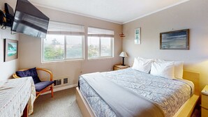 A hotel room with a bed, a chair, a table, a TV, and a window with blinds. - Oceanfront Nye Beach condo with gorgeous corner view & beach access (Newport)