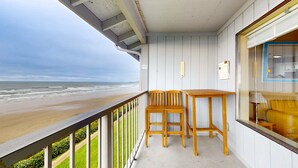 A balcony with a view of the beach, wooden furniture, and a window with blinds. - Oceanfront Nye Beach condo with gorgeous corner view & beach access (Newport)