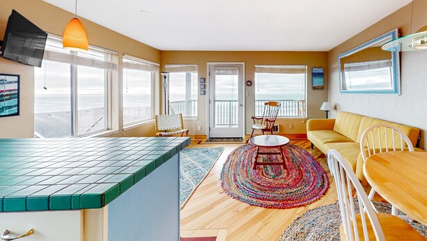 A spacious living area with a kitchen counter, dining table, and sofa, large windows, and a view of the sea. - Oceanfront Nye Beach condo with gorgeous corner view & beach access (Newport)