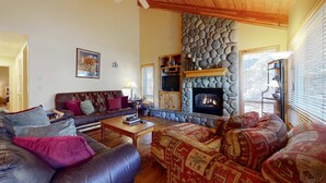 A cozy living room with a stone fireplace, leather sofas, a television, and a ceiling fan. - Sunriver home with WiFi, fireplace, private hot tub, & SHARC passes - dogs ok (Sunriver)