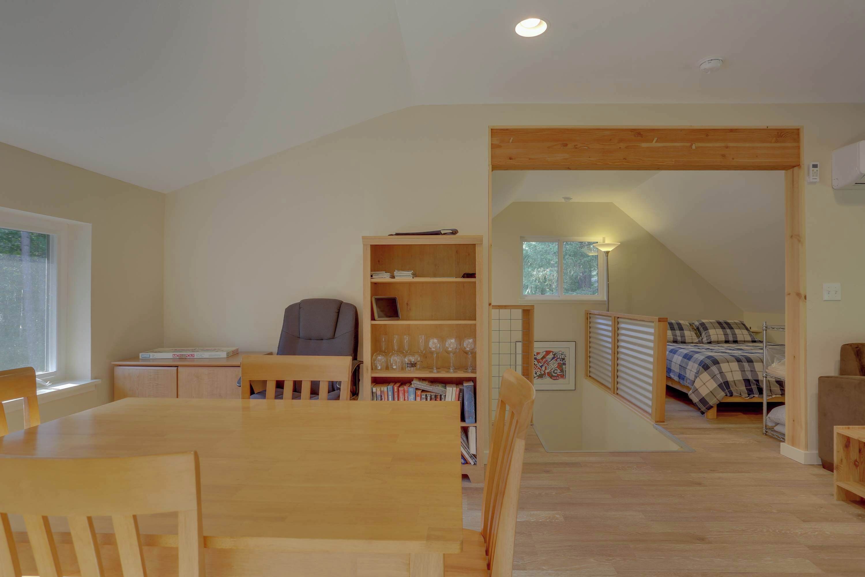 Una habitación con una mesa de comedor de madera, sillas, una estantería, una cama y una escalera que sube a un nivel superior.