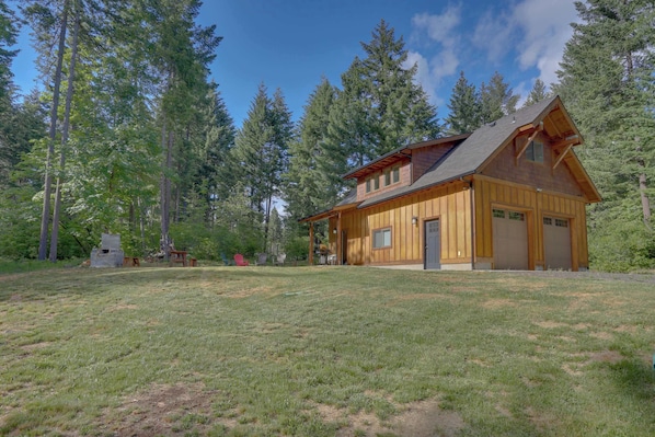 A wooden cabin with a garage, surrounded by a grassy yard and tall trees.