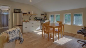 Una cocina con refrigerador, microondas, estufa y una mesa de comedor de madera con sillas. Hay ventanas con vistas al follaje.
