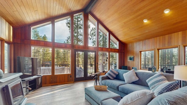 A spacious living area with a large window, a sofa, a television, and a grill. - Whimsical tree-lined stay with wood stove, AC, shared amenities, and large deck (Black Butte Ranch)