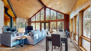 A spacious living area with a sofa, TV, and dining table, large windows, and wooden ceiling. - Whimsical tree-lined stay with wood stove, AC, shared amenities, and large deck (Black Butte Ranch)