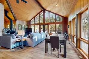 A spacious living area with a sofa, TV, and dining table, large windows, and wooden ceiling.