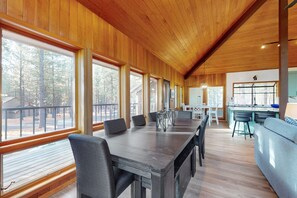 A spacious room with a long dining table, multiple chairs, and large windows offering a view of the forest.