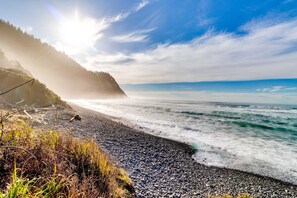 A coastal beach scene with a rocky shoreline, waves, and a forested cliff. - Charming home with ocean views, patio, BBQ grill, WiFi, W&D, walk to beach (Cannon Beach)