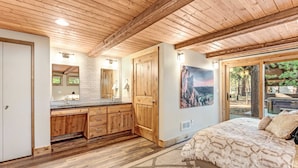 Ein Schlafzimmer mit einem großen Bett, Holzbalken an der Decke, einem Badezimmer mit Waschbecken und Spiegel sowie Blick ins Freie.