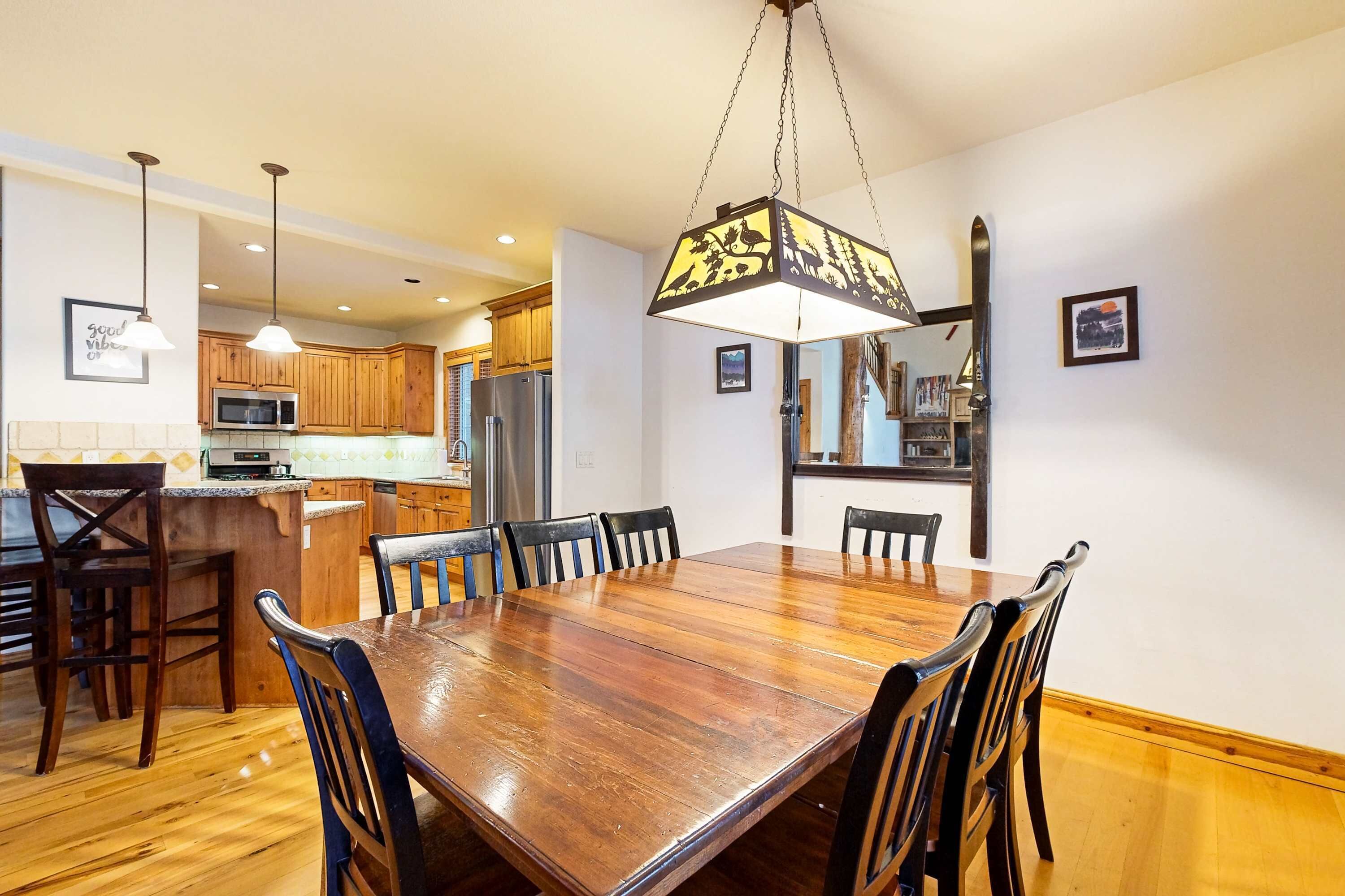A dining area with a wooden table and chairs, a hanging light fixture, wooden cabinets, and a kitchen in the background.