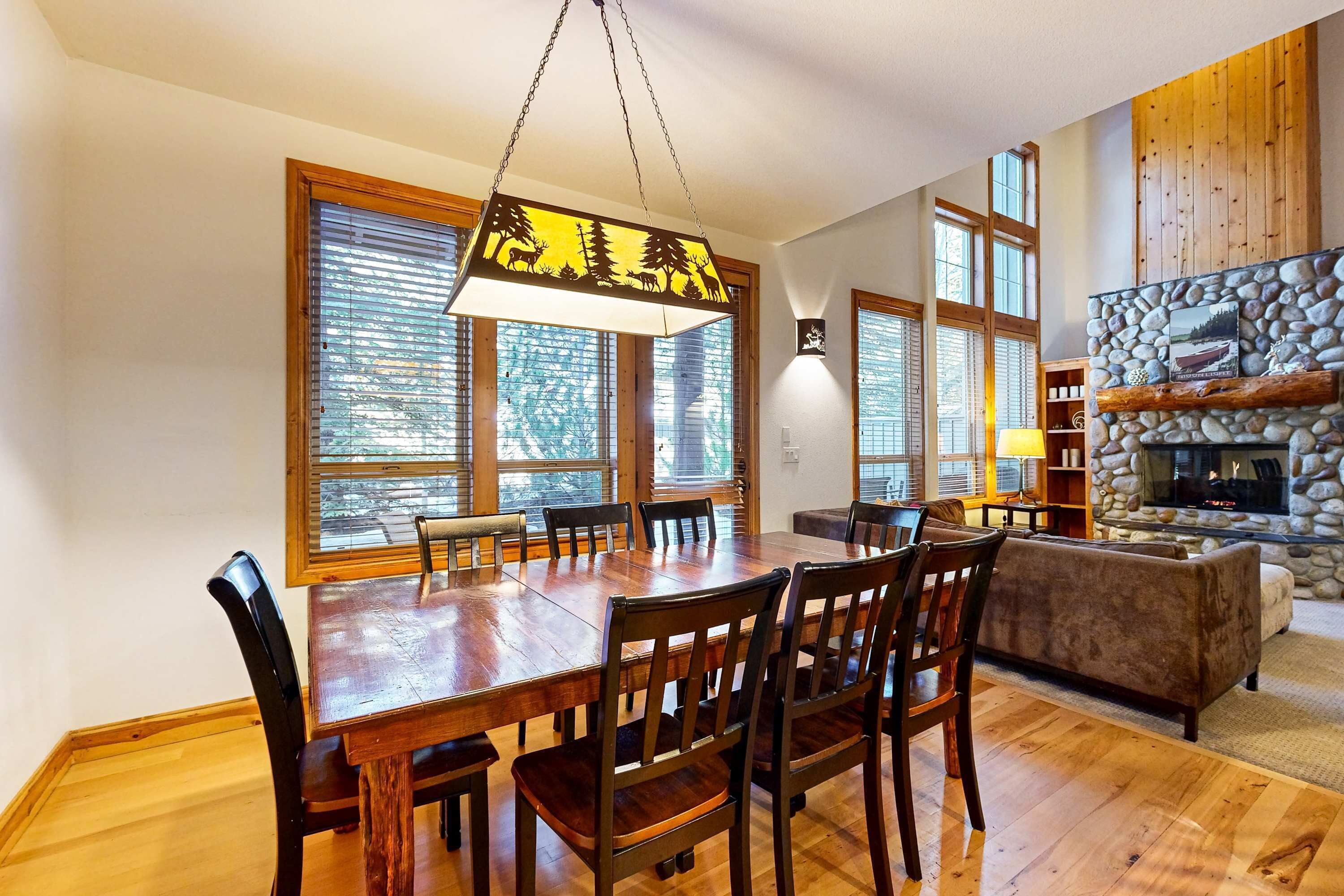 Comedor con mesa y sillas de madera, lámpara colgante, ventanas amplias y chimenea de piedra.