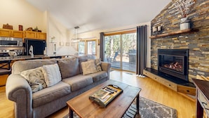 Ein modernes Wohnzimmer mit Sofa, Couchtisch, Kamin, Küchenbereich und großen Fenstern.