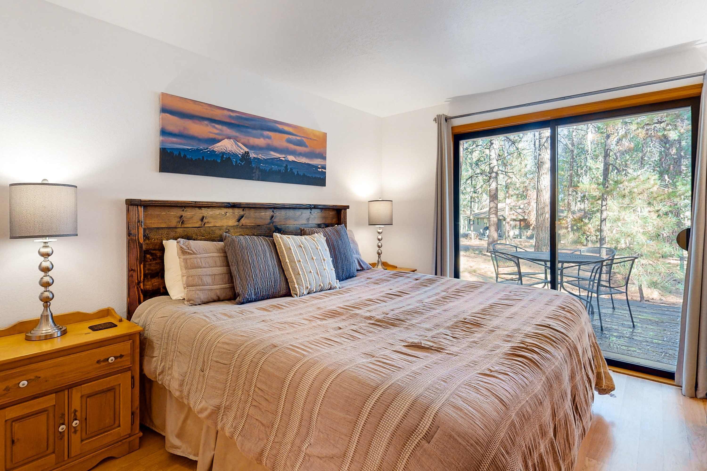 A bedroom with a large bed, a wooden headboard, two bedside lamps, a nightstand, and a view of the outdoors through a sliding glass door.