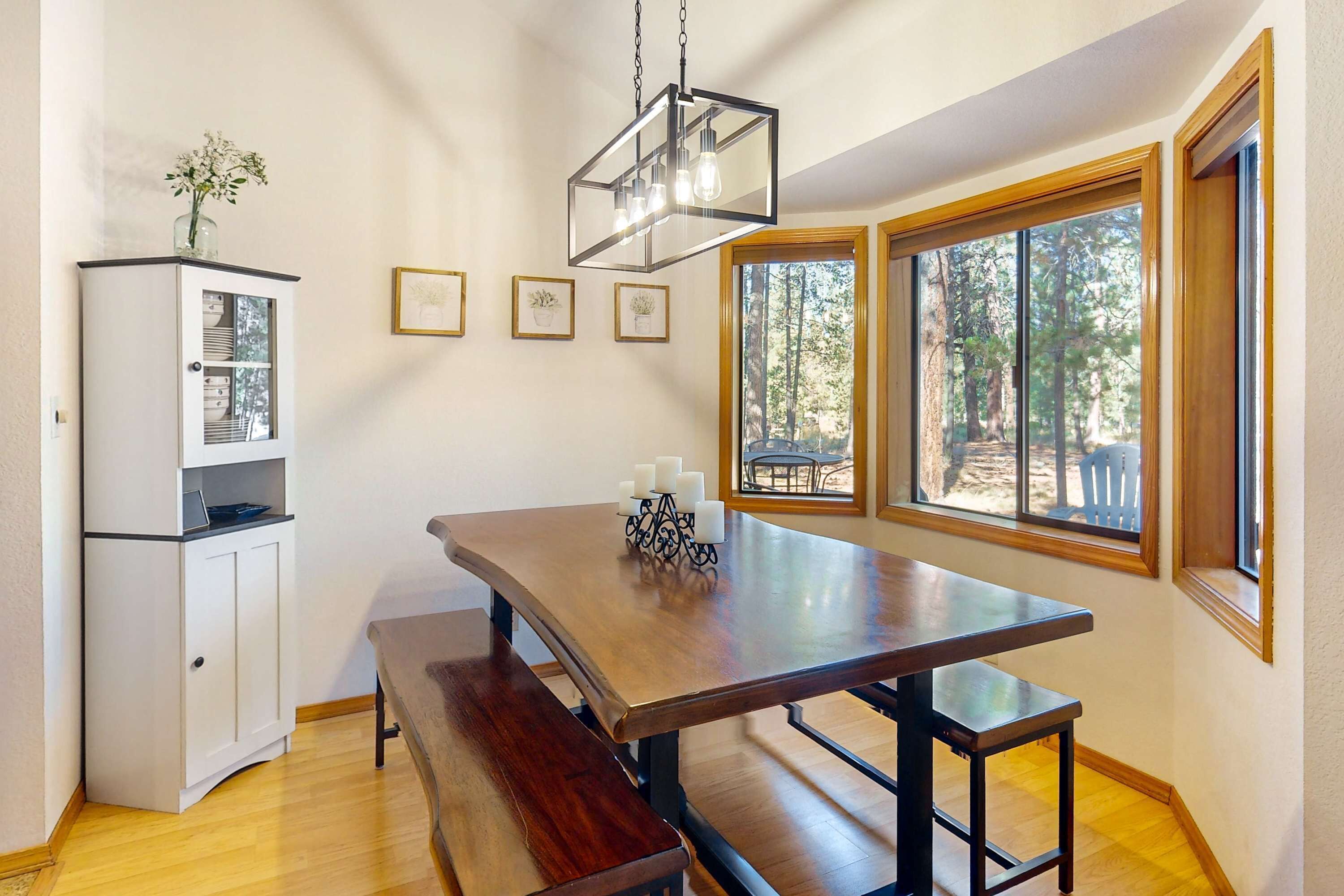 A dining area with a wooden table, benches, a hanging light fixture, and a view of trees through large windows.