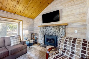 A cozy cabin interior with a stone fireplace, a flat-screen TV mounted above it, a sofa, and a window with a view of the outdoors. - Central Oregon cabin with SHARC amenities, fireplace, & grill - near skiing (Sunriver)