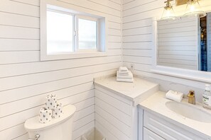 Ein Badezimmer mit weißem Holzvertäfelung, einer Toilette, einem Waschbecken mit Spiegel und einem Fenster.