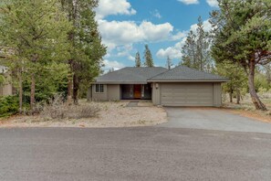 A single-story house with a garage, surrounded by pine trees, under a partly cloudy sky.