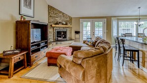 Ein Wohnzimmer mit Kamin aus Stein, Fernseher, einer Couch und einer Küchenzeile mit Bar.
