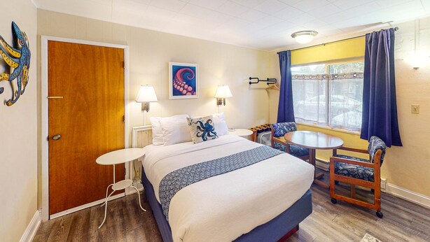 Una habitació d'hotel amb llit, taulella de nit, cadira, làmpada, obra d'art i finestra amb cortines.