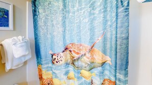 Un bany amb una cortina de dutxa que representa una tortuga marina i peixos, un penjador de tovalloles amb una tovallola blanca i un quadre al mur.