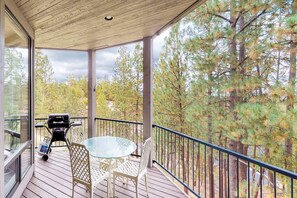 Terraza exterior techada con mesa y sillas de vidrio, una parrilla y vista a un bosque.