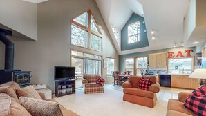 A spacious living room with a fireplace, large windows, a kitchen area, and a dining table. - Beautiful & spacious home with a private hot tub, fast WiFi, AC, & SHARC access (Sunriver)
