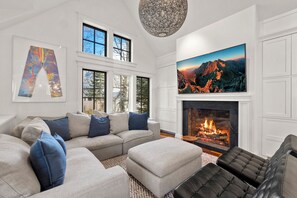 Un salon comprenant un canapé d’angle, un pouf, une cheminée et un grand tableau représentant un paysage de montagne.