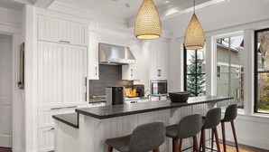 Una cucina moderna con mobili bianchi, un'isola centrale con sgabelli da bar, illuminazione a sospensione e vista su zona residenziale.
