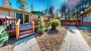 A wooden deck leading to a house with a sliding glass door, surrounded by a fenced yard with trees and a clear sky. - Cozy forest retreat near trails, slopes & Sunriver - perfect for year-round fun! (Bend)