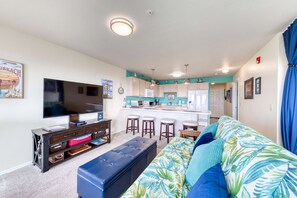 A living room with a TV, sofa, ottoman, and kitchen area with bar stools.