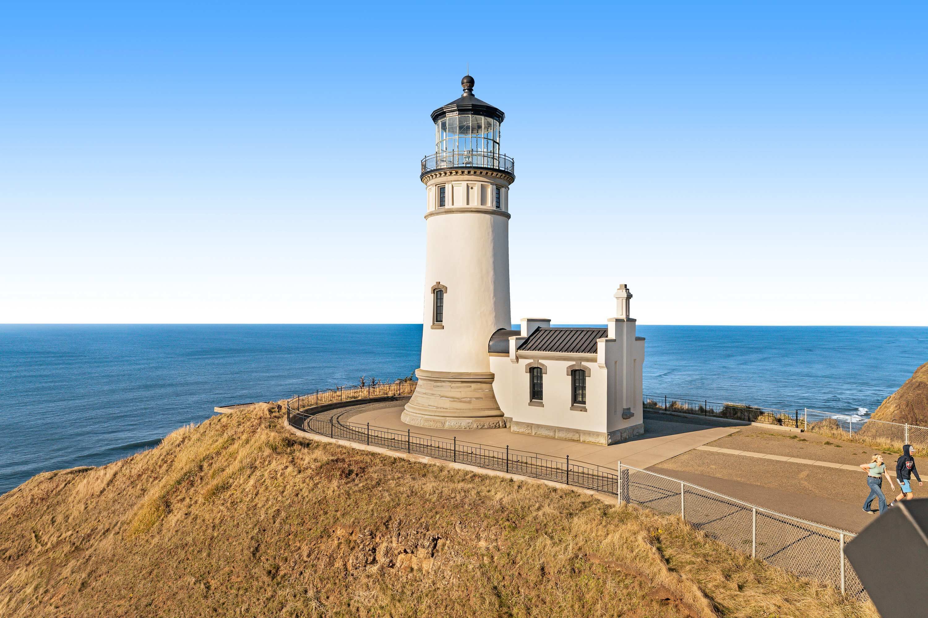 A lighthouse with a circular base, a smaller attached structure, and a paved walkway leading to it.