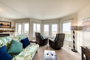A living room with a sofa, two armchairs, a coffee table, and a floor lamp. Large windows provide a view of the outdoors. - Dog-friendly oceanfront condo with electric fireplace - walk to the beach (Long Beach)