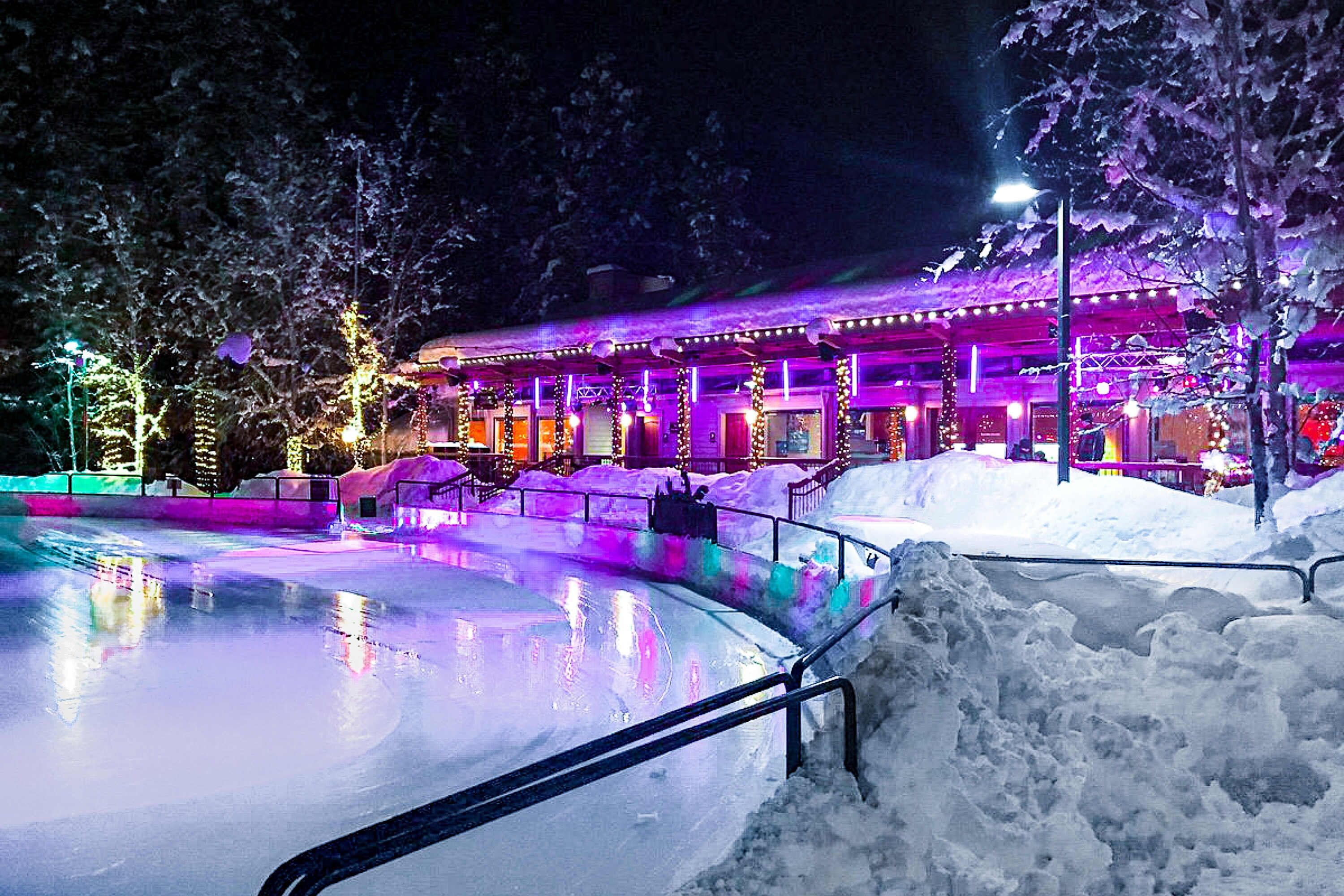 Uma área externa coberta de neve, com piscina, guarda-corpos e um edifício iluminado em roxo.