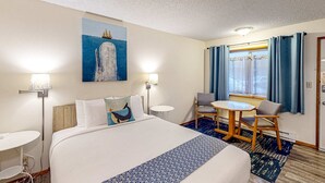 Une chambre d’hôtel avec un lit, une petite table, deux chaises, une lampe, un tableau et une fenêtre avec des rideaux.