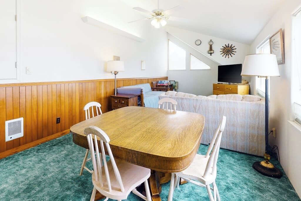 Een kamer met een houten eettafel, witte stoelen, een bed met een blauw hoofdeinde, een televisie en een plafondventilator.