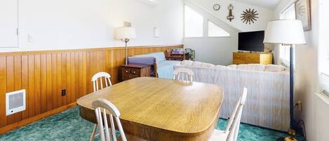Een kamer met een houten eettafel, witte stoelen, een bed met een blauw hoofdeinde, een televisie en een plafondventilator.