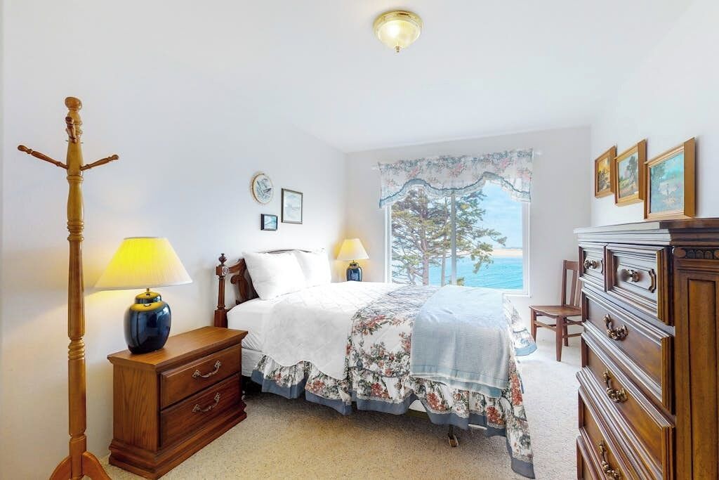 Ein Schlafzimmer mit einem Bett, Nachttischen, einem Schminktisch, einer Lampe, einem Fenster mit Aussicht und einer Deckenleuchte.