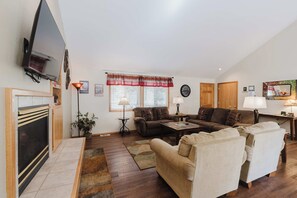 Ein Wohnzimmer mit Kamin, einem an der Wand befestigten Fernseher, einer Sofagarnitur, einem Couchtisch und einem Esstisch mit Stühlen.