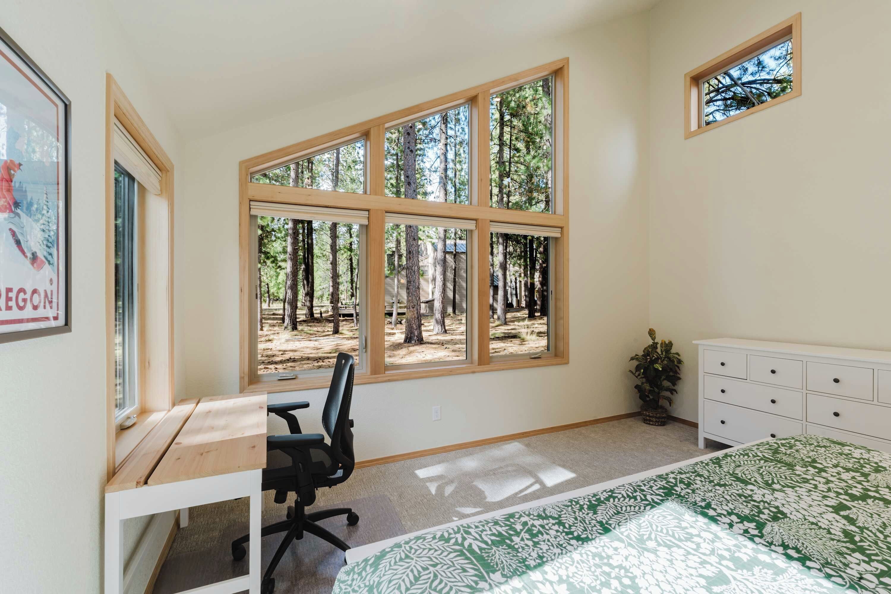 A bedroom with a large window, a desk, a chair, a dresser, and a small potted plant.