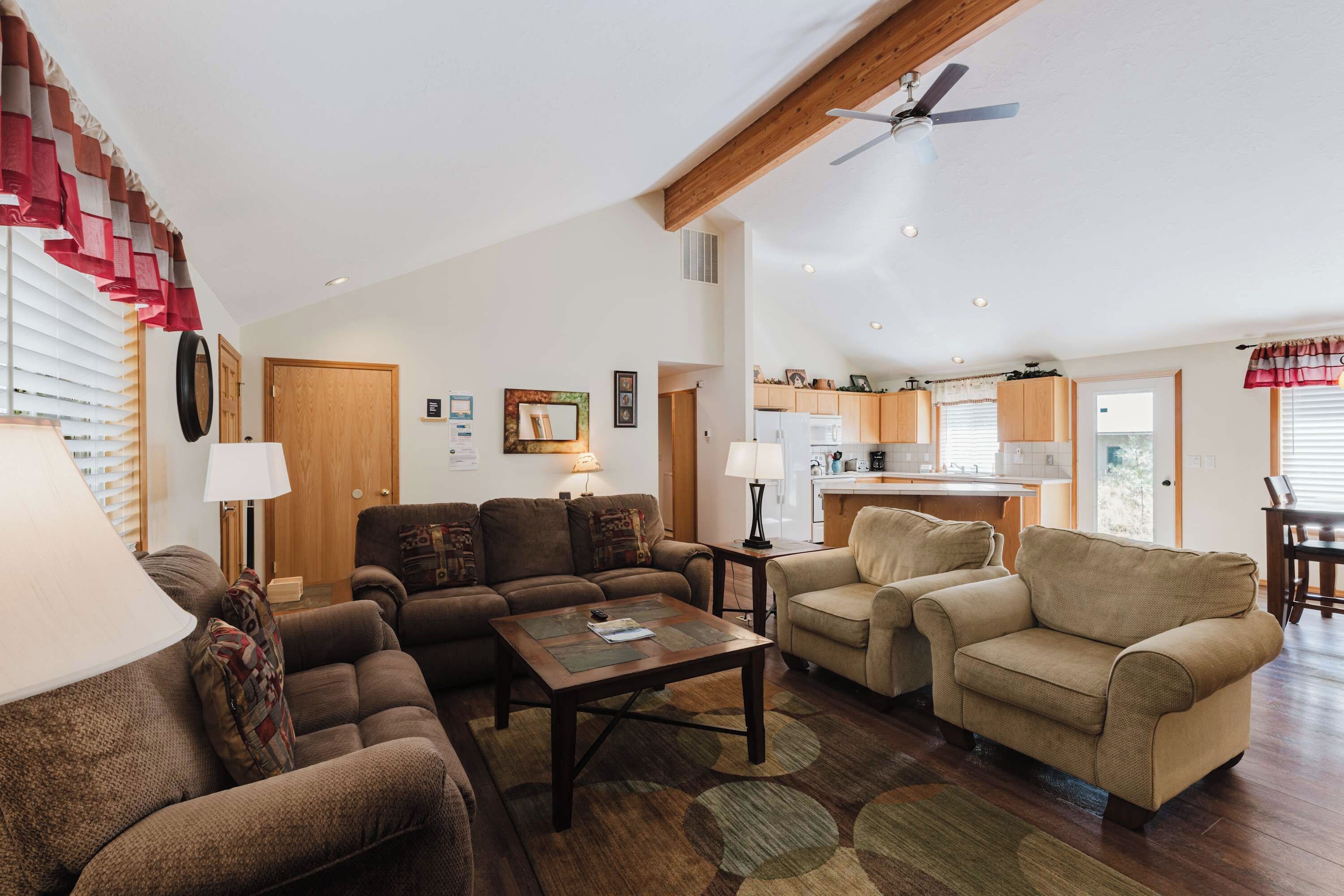 A spacious living room with a high ceiling, a ceiling fan, and a kitchen area with wooden cabinets and a window.