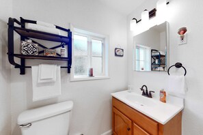 A bathroom with a white toilet, a wooden vanity with a mirror, a shelf with towels, and a window with frosted glass.