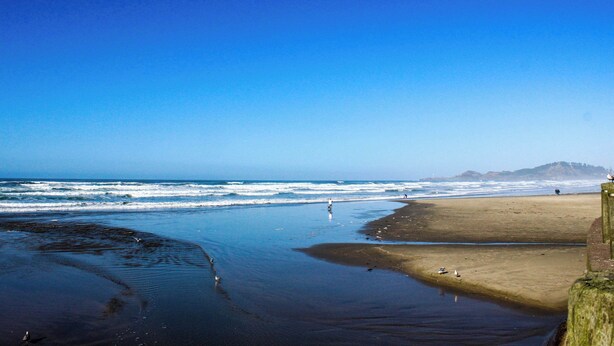 Una playa con olas, orilla arenosa y una montaña a lo lejos.