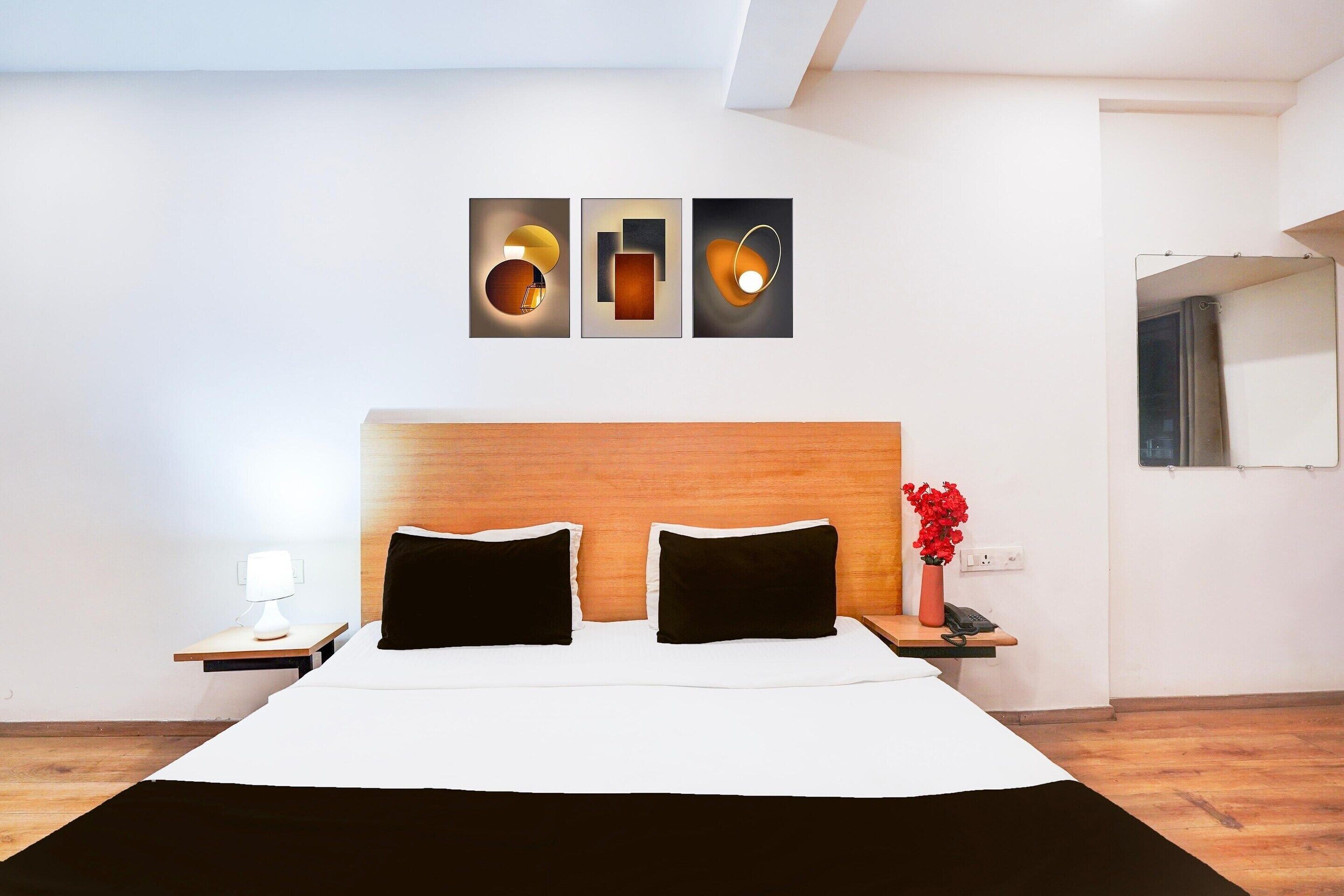 A hotel room with a wooden headboard, a bed with black and white bedding, a nightstand with a lamp, a vase with red flowers, and three wall-mounted artworks.