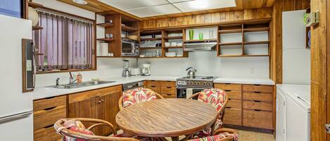 Een kleine keuken met houten keukenkastjes, een eettafel met stoelen en een koelkast.