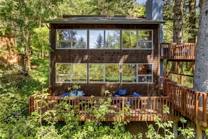 Uma cabana de madeira de dois andares, com janelas amplas, um deck com assentos e um deck de casa na árvore.